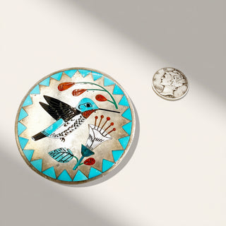 Decorative circular brooch with bird design next to a coin on a light gray background