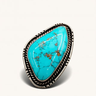 A sterling silver ring featuring a large, irregularly shaped turquoise stone with natural brown matrix veining, set in a detailed rope-textured bezel on a light cream background.