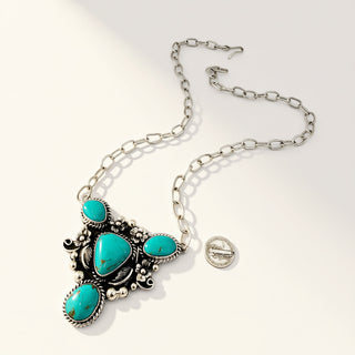A silver-toned necklace with a Southwestern-style pendant featuring four turquoise-colored stones, displayed on a light beige background next to a US quarter coin for scale.