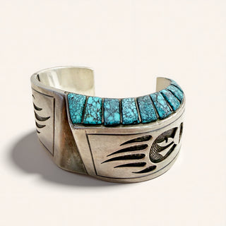 A wide sterling silver cuff bracelet featuring a row of turquoise stone inlays on the front, photographed on a clean off-white background.