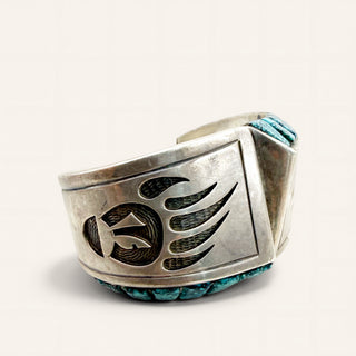 A wide silver cuff bracelet featuring a large engraved winged emblem on the front and turquoise-colored braided accents along the top and bottom edges, photographed on a clean off-white background.