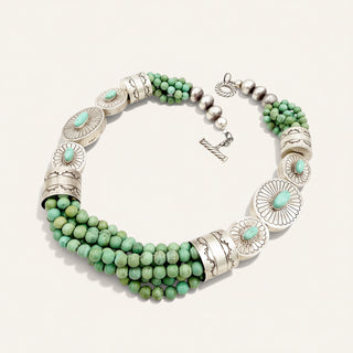 A multi-strand turquoise and silver statement necklace displayed on a clean, light beige background. The necklace features multiple strands of round green turquoise beads, textured silver cylindrical beads, and larger silver disc elements, each decorated with a small round turquoise cabochon.