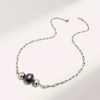 A silver chain necklace featuring three polished metallic beads, including one larger central bead with a dark reflective center, displayed on a clean white background.
