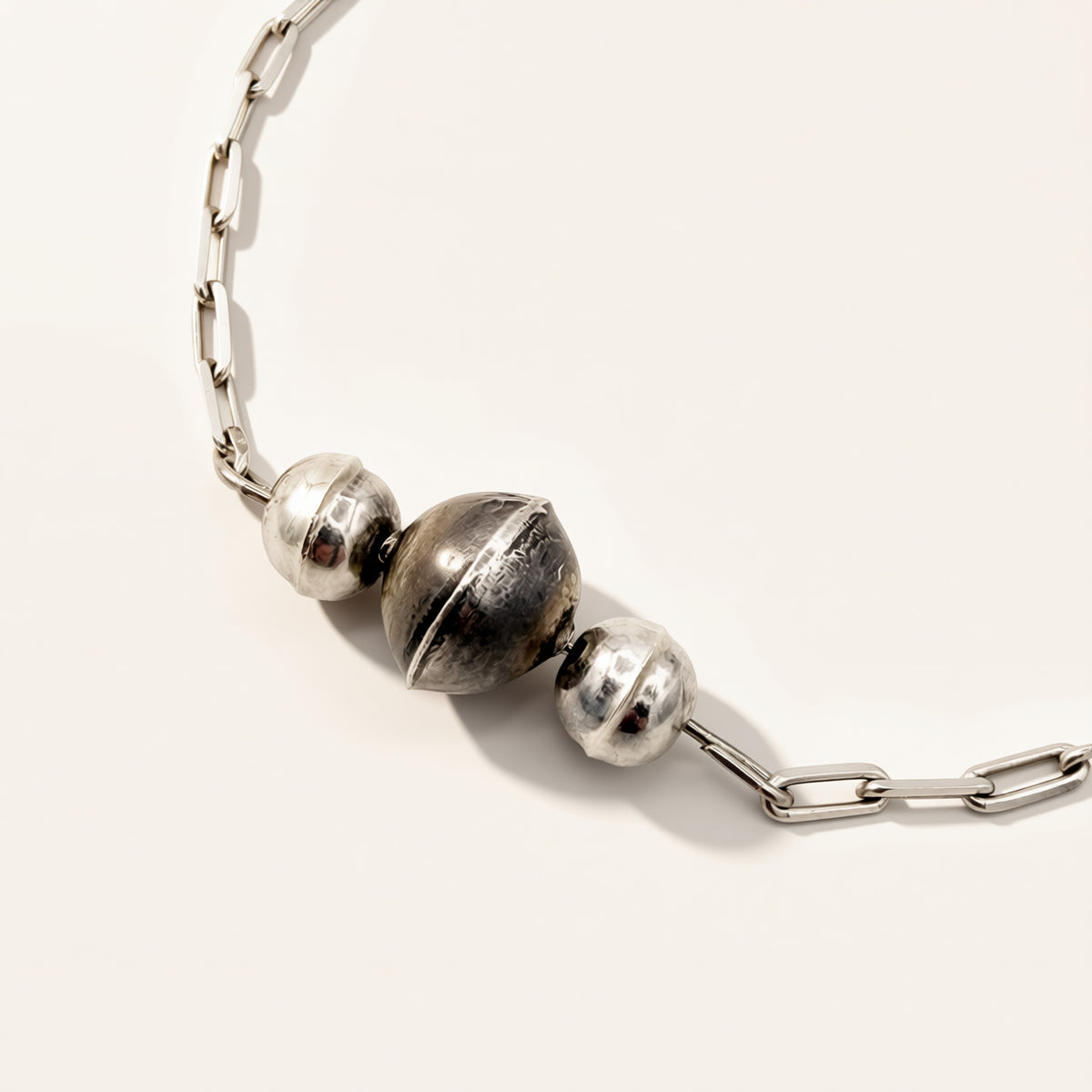 A close-up of a silver chain necklace featuring three metallic spherical beads, with one larger textured bead in the center and two smaller polished beads on either side, set against a plain off-white background.