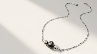 A silver chain necklace with three spherical beads, featuring a central dark metallic bead flanked by two polished silver beads, displayed on a light surface with soft diagonal shadows.