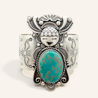 A wide sterling silver cuff bracelet featuring a large oval turquoise stone and a stylized face motif on the front, photographed against a plain light beige background.