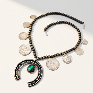 A silver-toned beaded necklace with a large open crescent pendant featuring a turquoise stone, displayed on a light beige background with several silver coins arranged nearby.