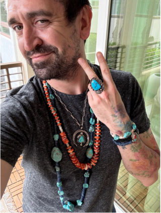 Man wearing multiple necklaces and rings with turquoise stones indoors.
