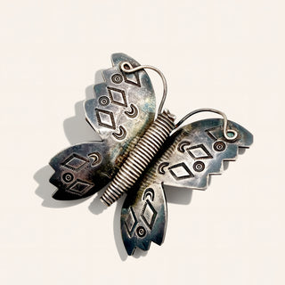 A butterfly-shaped brooch made of oxidized silver metal with engraved geometric patterns and a coiled spring-like center, photographed on a plain light background.