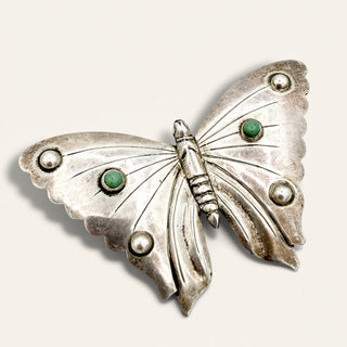 A vintage-style butterfly brooch made of silver-toned metal with small green stone accents and pearl-like details, shown on a plain light background.