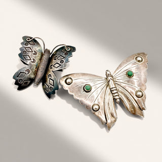 Two vintage-style butterfly brooches on a light gray background. One brooch is dark silver with blackened detailing, and the other is silver with green stone accents and raised circular details on the wings.