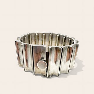 A wide, chunky silver metal cuff bracelet with a segmented, flared design and a circular clasp, photographed on a light beige background.