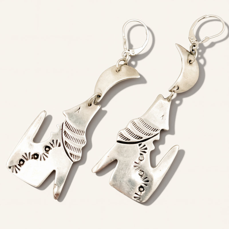 A pair of sterling silver dangle earrings shaped like stylized cat heads, featuring textured details and circular ear loops, displayed on a light cream background.
