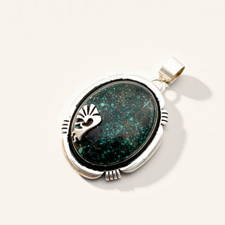 A sterling silver oval pendant featuring a dark green and turquoise inlay with a silver sunburst design, shown on a light cream background.
