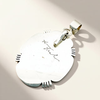 A sterling silver pendant with a polished, rounded organic shape and a bail at the top, featuring a small hand-written style script engraving in the center. The pendant is photographed on a warm beige background with soft shadows.