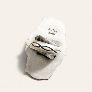 A silver ring with a black infinity symbol, displayed on a textured white shell stand with 'R. Tom Sterling' printed on it, set against a warm off-white background.