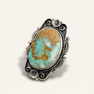 An ornate sterling silver ring featuring a large oval turquoise stone with prominent brown matrix, set against a cream background.