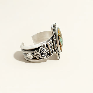 Silver cuff with intricate designs and a colorful gemstone on a light background