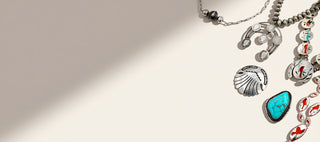 A styled flat-lay of silver-toned jewelry charms and beaded chains on a warm cream background with a diagonal shadow. The jewelry includes a seashell charm, a turquoise stone charm, and other decorative charms.