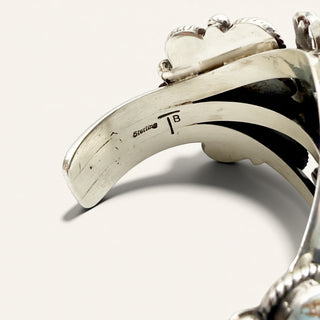 Close-up of a sterling silver cuff bracelet showing the interior engraving with ' sterling TB' and polished metal details on a light beige background.