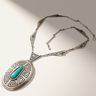 A Southwestern-style sterling silver pendant necklace with a large oval medallion featuring a turquoise inlay, set against a light beige background with soft shadows.