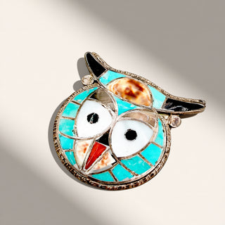 An owl-shaped brooch featuring turquoise inlay, white enamel eyes, a red beak, and black ear tips, set in silver-toned metal with a textured border, photographed on a light beige background with soft diagonal shadows.