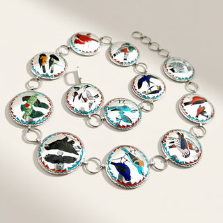 A silver-toned chain bracelet featuring multiple oval inlay charms decorated with colorful bird illustrations on a white background, arranged on a light beige surface.