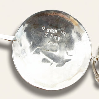 Close-up of the polished silver back of a round pendant or locket, showing engraved maker's marks reading 'Q QUAM SR ZUNI' on a light neutral background.
