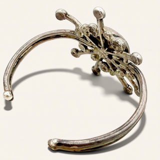 A silver-toned open cuff bracelet featuring an intricate, textured floral or starburst cluster at the top, photographed on a light beige background.