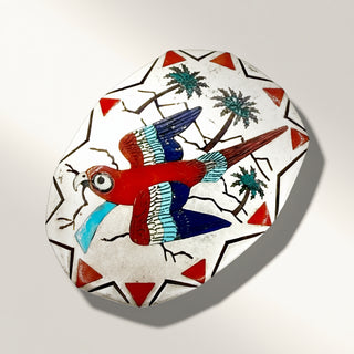 A hand-painted ceramic plaque featuring a red and blue parrot, green palm trees, and red triangles on a white background with black geometric borders.