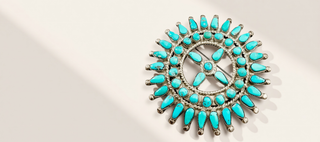 A circular silver-toned brooch or pin featuring multiple turquoise-colored stone accents arranged in a sunburst pattern, photographed on a clean off-white background with soft diagonal shadows.