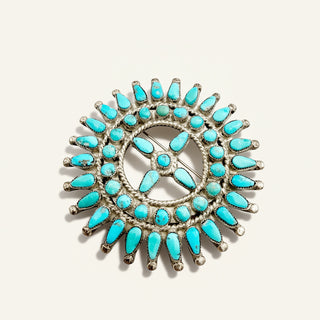 A circular silver-toned brooch featuring multiple turquoise-colored stones arranged in a sunburst pattern, photographed on a light beige background.