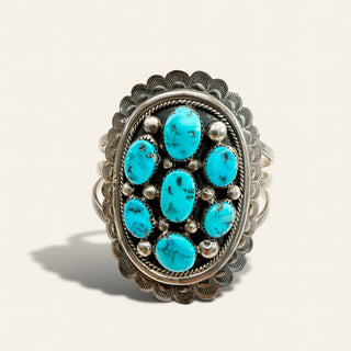 A silver ring featuring a cluster of seven oval turquoise stones in the center, surrounded by small silver beads and set within a scalloped silver bezel, shown on a light beige background.