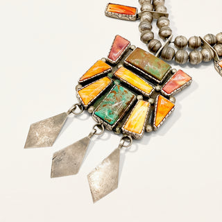 A close-up of a Southwestern-style silver necklace featuring rectangular and triangular turquoise, orange, and red stones, with three silver diamond-shaped dangles hanging from the center, set against a plain off-white background.