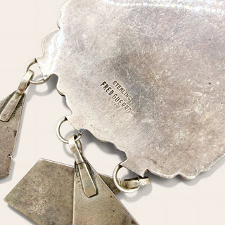 Close-up of a sterling silver pendant necklace with a scalloped triangular pendant and dangling geometric elements, showing the engraved 'STERLING FRED GUERRO' mark on a light beige background.