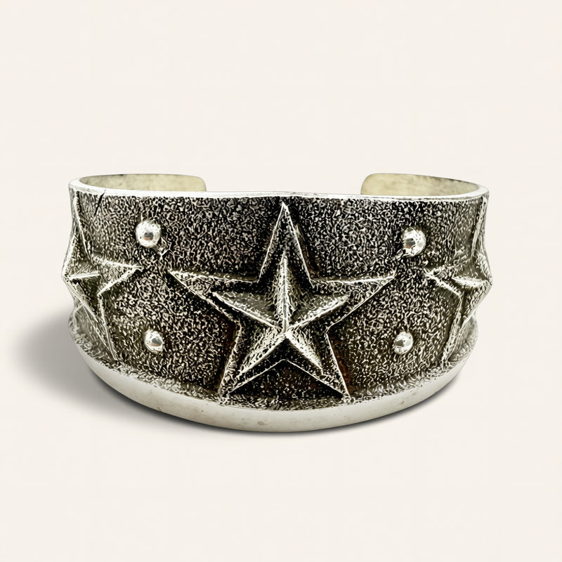 A wide silver cuff bracelet featuring a textured star design with small silver studs, photographed on a light beige background.