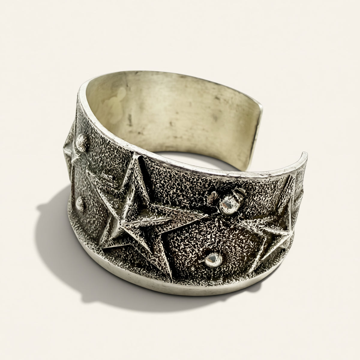 A wide silver cuff bracelet featuring a textured star pattern and small clear rhinestone accents, photographed on a light beige background.