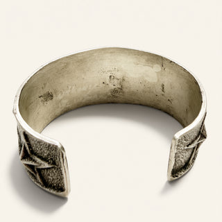A wide silver cuff bracelet with textured geometric star-like details on the open ends, displayed on a light beige background.