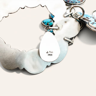 A close-up of a silver-toned jewelry piece with a scalloped shell-shaped border, turquoise stone accents, and a white teardrop charm stamped with 'J. Tso'.