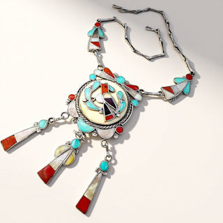 A Southwestern-style silver necklace featuring a large oval pendant and dangling tassel charms, decorated with turquoise, red, white, and yellow inlay stones on a clean white background.