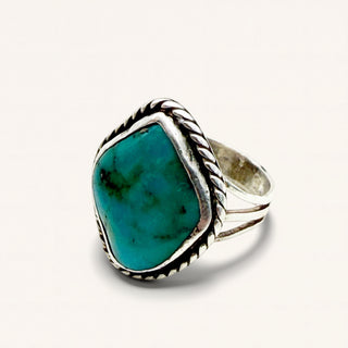 A sterling silver ring featuring a large, irregular-shaped turquoise stone with natural green and blue marbling, set in a textured bezel with a twisted rope border and a multi-pronged band, shown on a plain off-white background.
