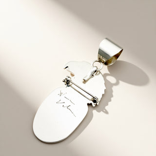 A silver pendant, featuring a safety pin clasp and a small signature detail, shown on a warm beige background with soft diagonal shadows.
