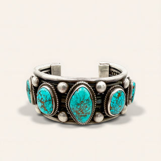 A Southwestern-style cuff bracelet featuring four large turquoise stones and four smaller turquoise stones, set in silver with silver beads and rosettes, on a light beige background.