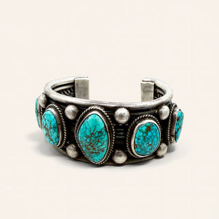 A sterling silver cuff bracelet featuring five oval turquoise stones with dark matrix, accented by small round silver beads, photographed on a light beige background.