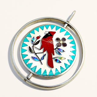 A circular silver-toned pendant brooch featuring a red cardinal bird and turquoise, white, and iridescent mosaic details on a light cream background.