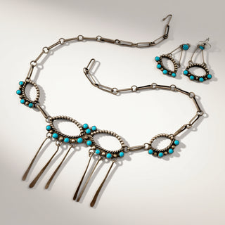 A silver-toned jewelry set featuring a necklace and matching earrings with turquoise blue bead accents and dangling metal elements, displayed on a light gray background.
