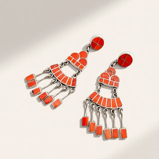A pair of statement dangle earrings with bright orange-red enamel inlay and silver-tone metal, displayed on a clean off-white background with soft diagonal shadows.