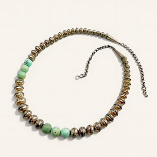 A beaded necklace featuring metallic bronze and gold-toned disc beads with a row of light green round beads, displayed on a light cream background.