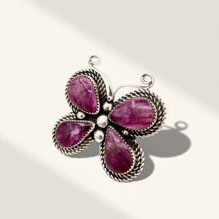 A butterfly-shaped silver pendant featuring four teardrop-shaped deep pinkish-purple gemstones and small silver beads at the center, shown on a light beige background with soft shadows.