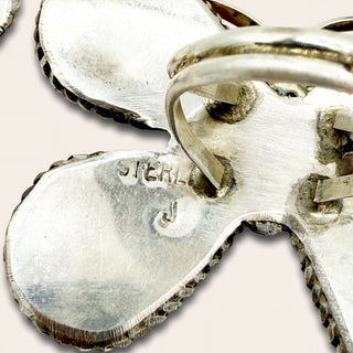 A close-up of a sterling silver ring,  showing the polished metal surface and engraved 'STERLING' text on one petal against a light beige background.